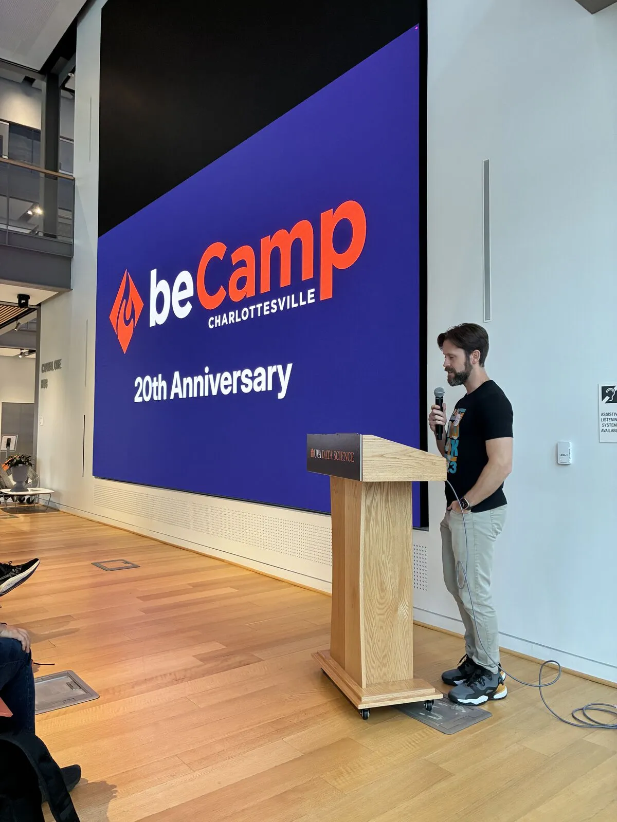 A speaker at the lectern beside the screen reading 'beCamp Charlottesville 20th Anniversary'