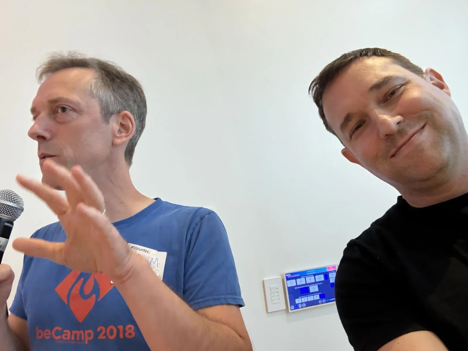 An organizer's selfie with a pitcher in a beCamp 2018 T-shirt, mid-gesture at the microphone