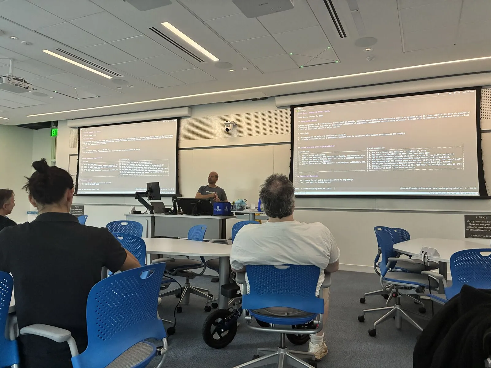 A session in a lecture room, a presenter at the front between two projected screens of text