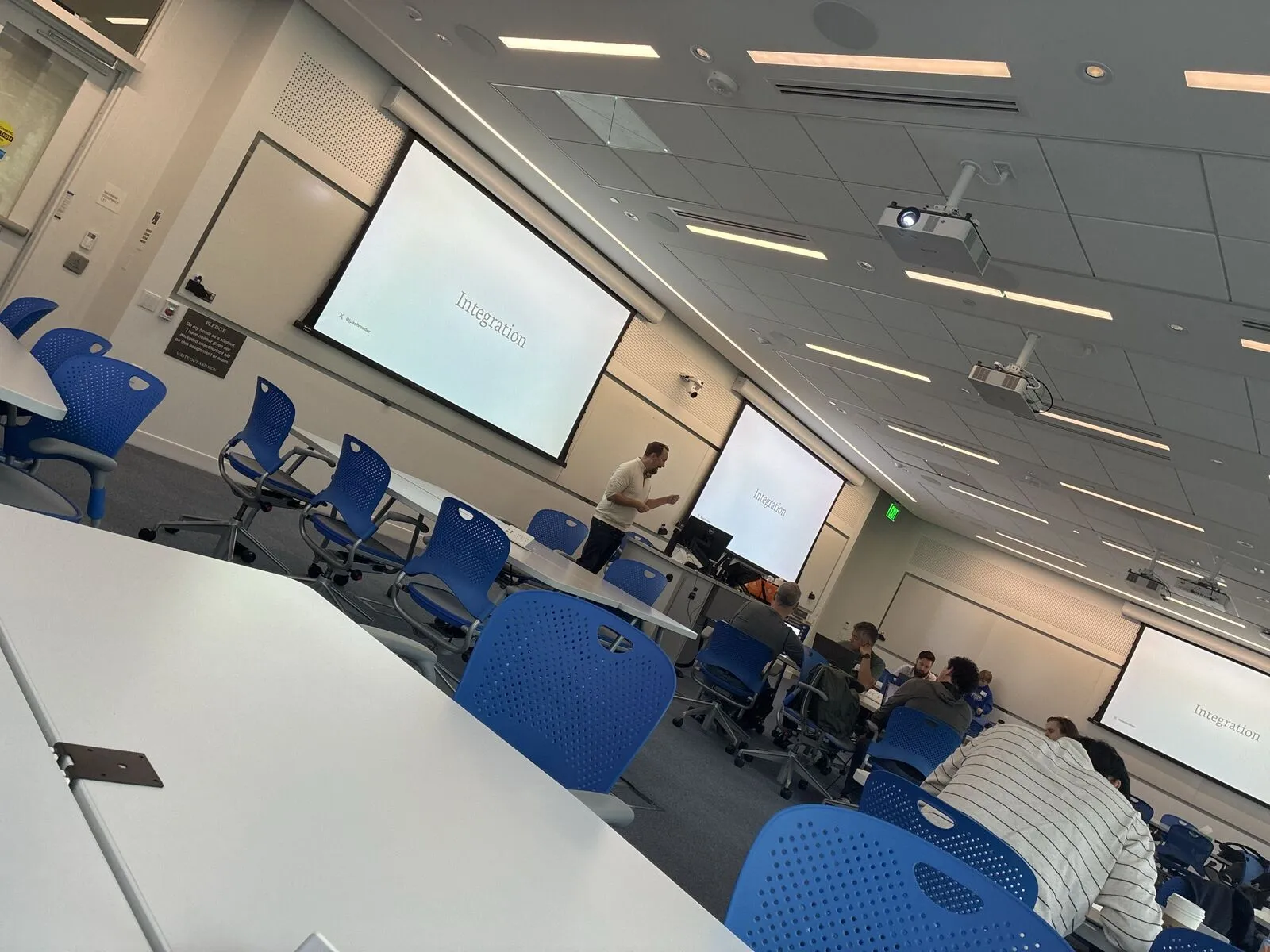 A lecture room session seen from the side, attendees in blue chairs facing the projected screens