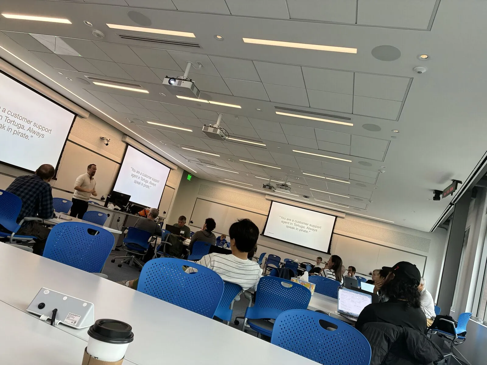 Attendees at desks watching a presenter in a lecture room, a coffee cup in the foreground