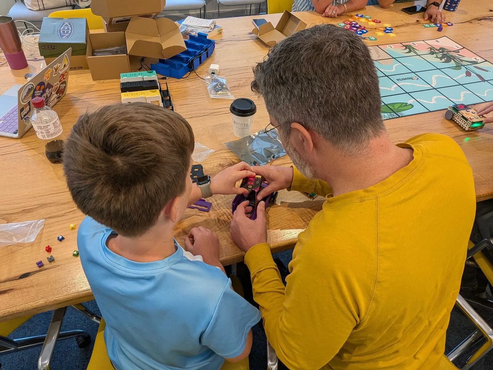 An adult and a child building a small robot together at a table covered in parts