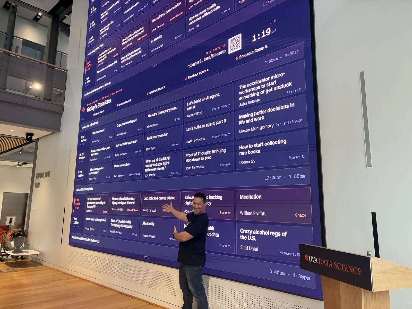 An organizer gesturing at the day's session board on the main hall's video wall