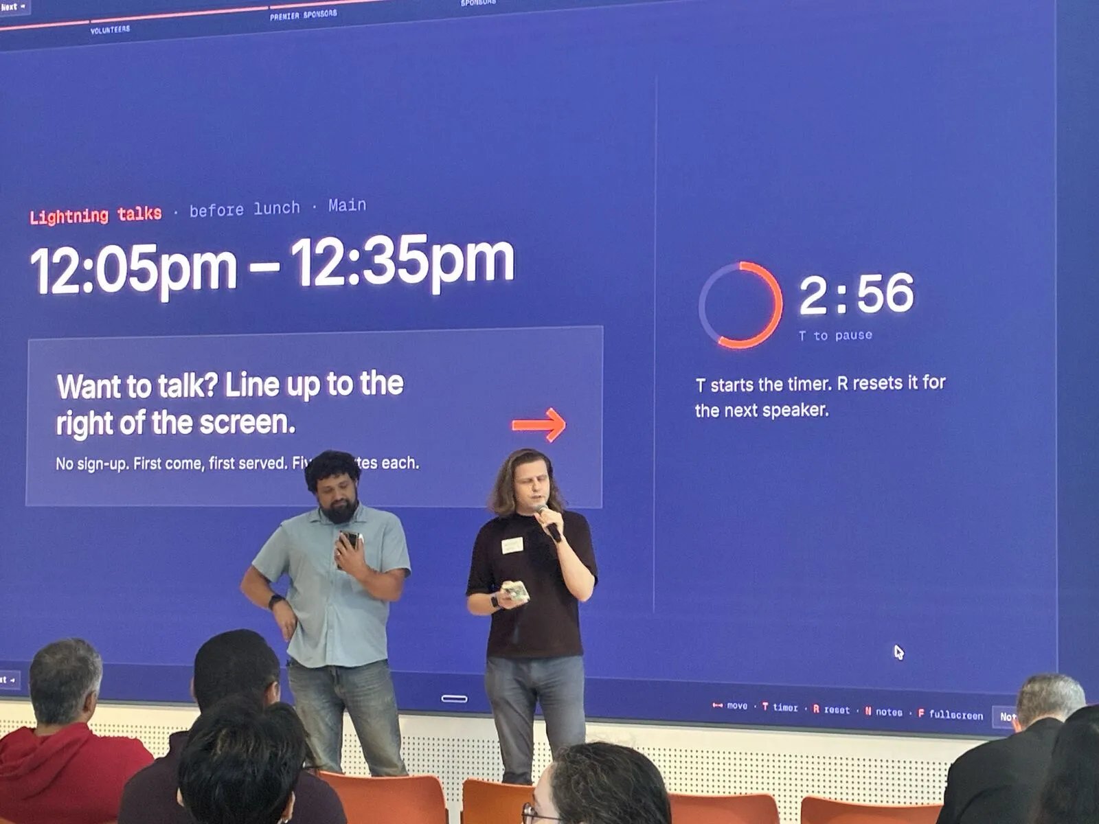 Two speakers giving a lightning talk in front of the video wall timer