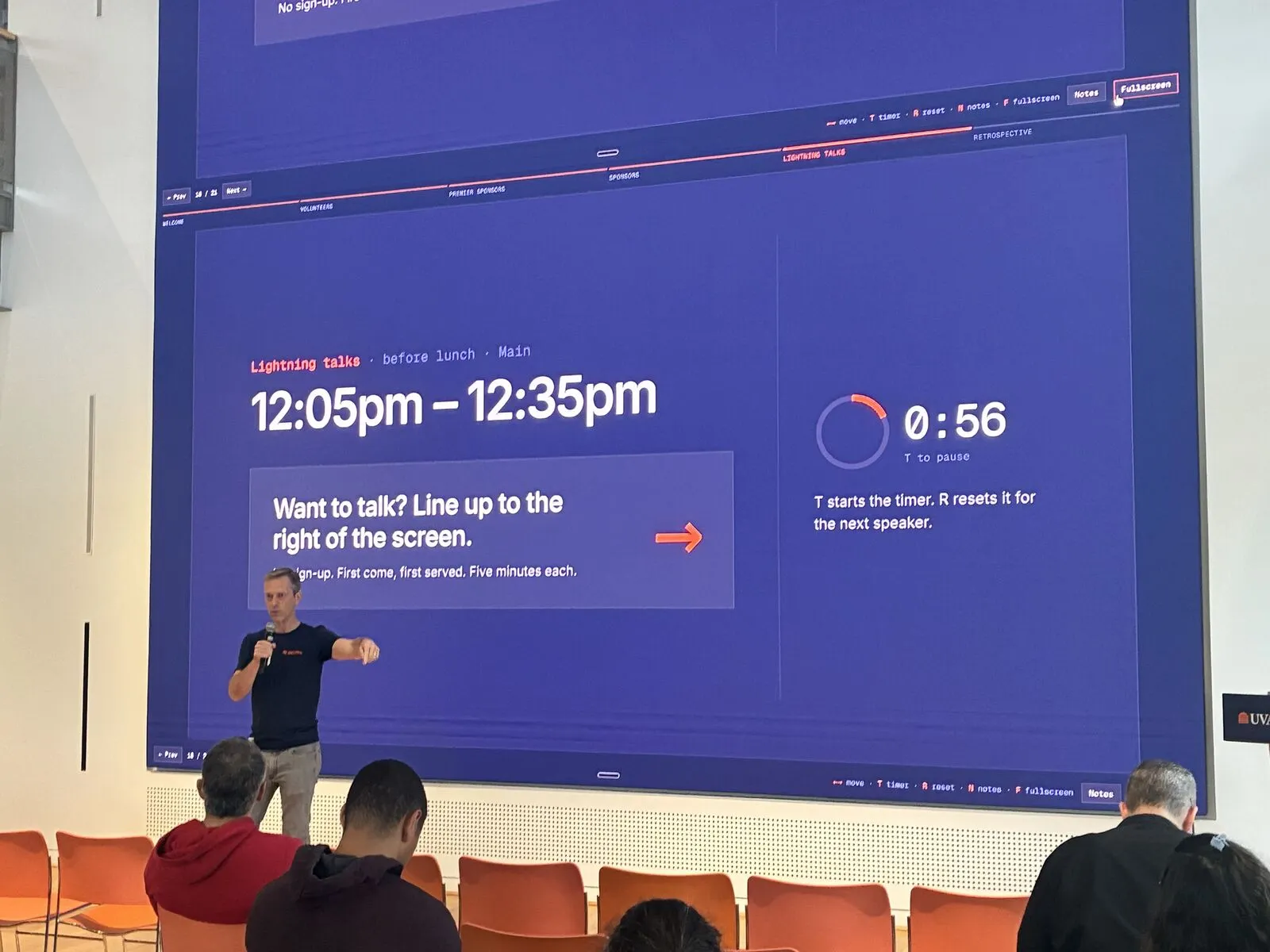 A lightning talk speaker under the video wall reading "Want to talk? Line up to the right of the screen"