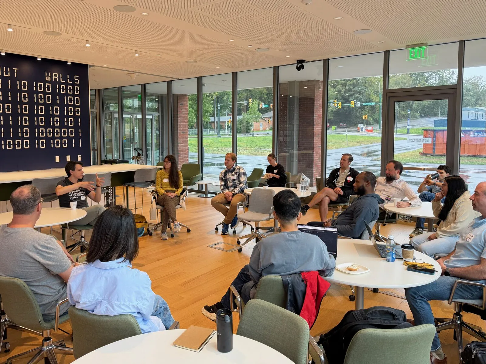 A discussion circle in a bright room with floor-to-ceiling windows and a wall of binary code