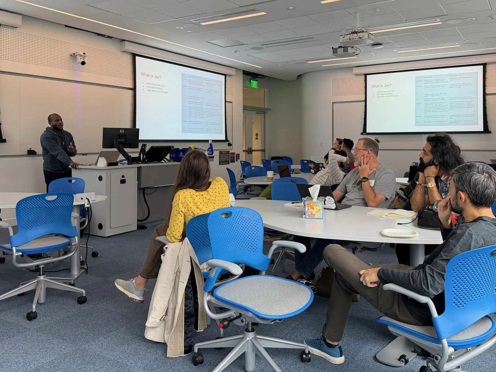 A presenter standing at the front of a lecture room, attendees in blue chairs around tables