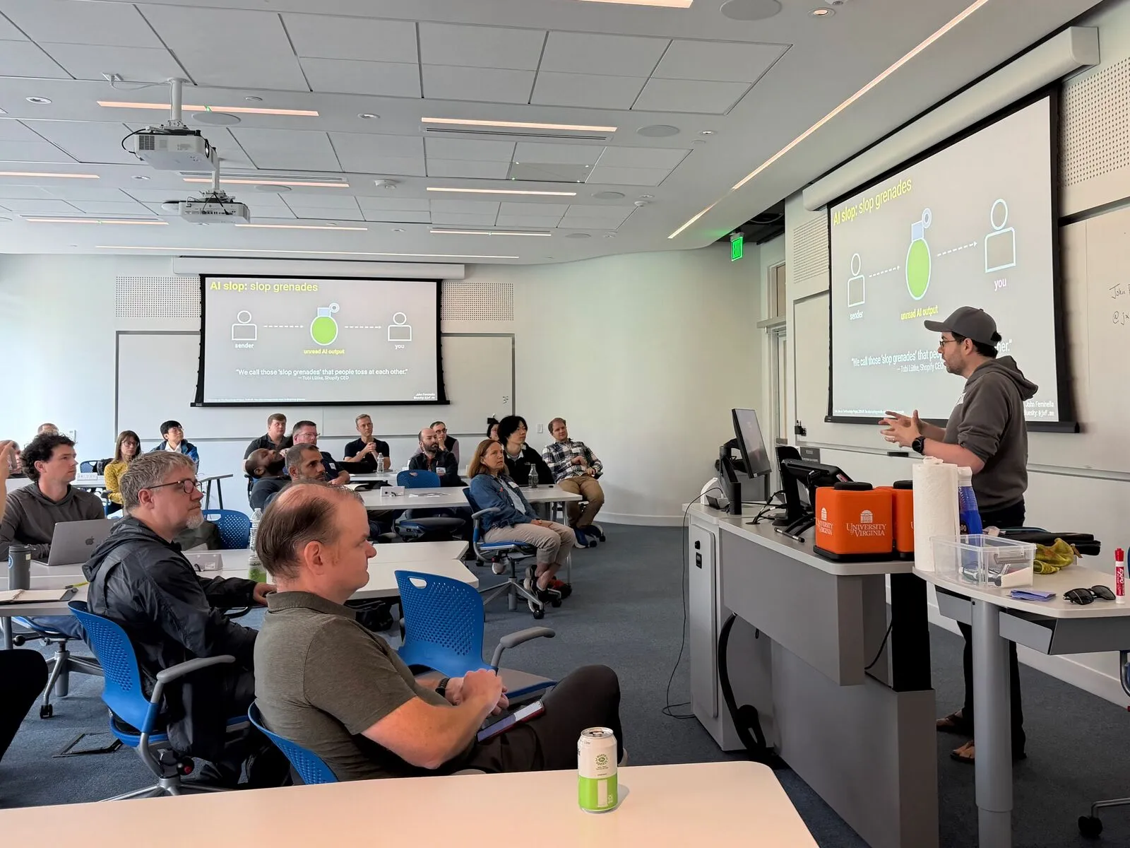 A presenter beside a projected diagram at the front of a lecture room, attendees listening