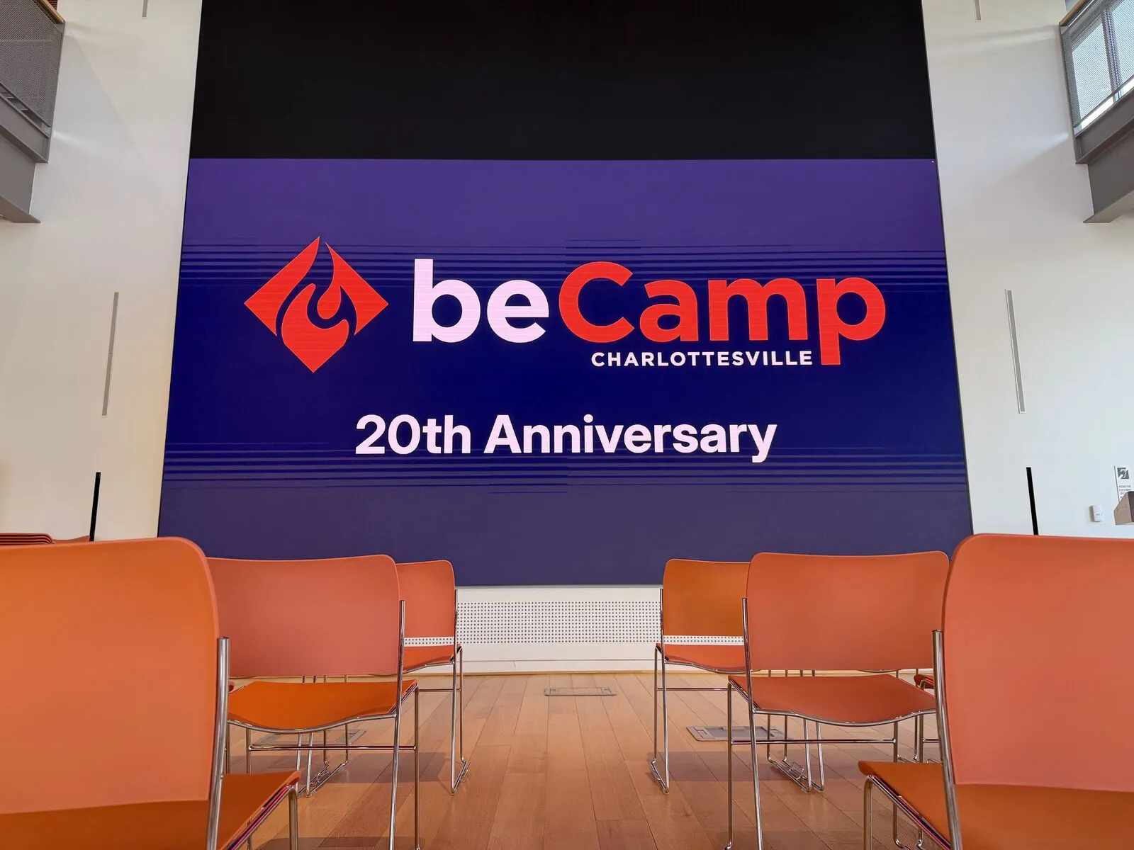 The empty stage before doors, the screen reading 'beCamp Charlottesville 20th Anniversary' over rows of orange chairs